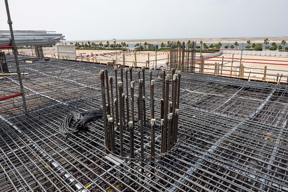 Beach Walk Iii By Imtiaz Construction site view showing steel rebar and framework for concrete, with a coastal landscape in the background.